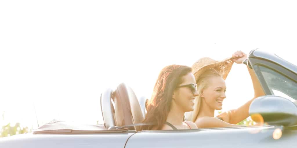 two girls in convertible driving away with lots of sunshine in sky