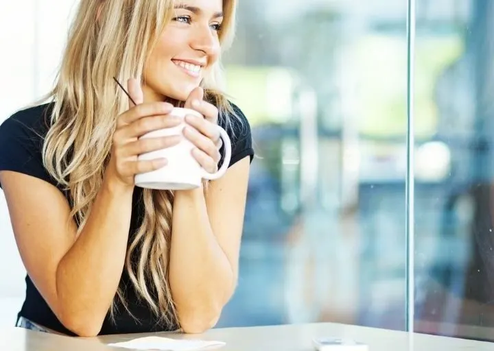 young woman with coffee, smiling, looking out of window at desk thinking about why do people want to be rich