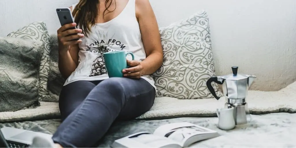 woman sitting on bed, reading on phone, with turquoise mug