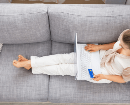 young woman laying on couch with laptop and credit card, wondering how to hide money from herself to stop spending it