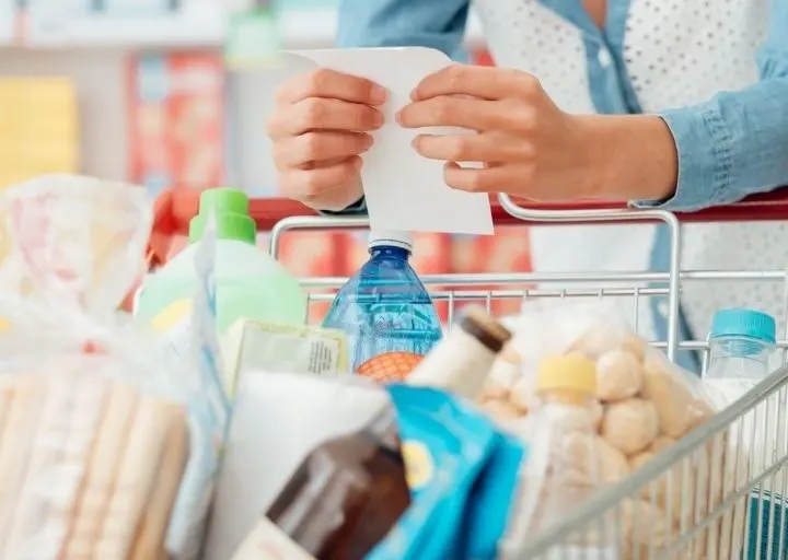 woman holding onto list and coupons at grocery store, with grocery cart, wondering about coupon fraud