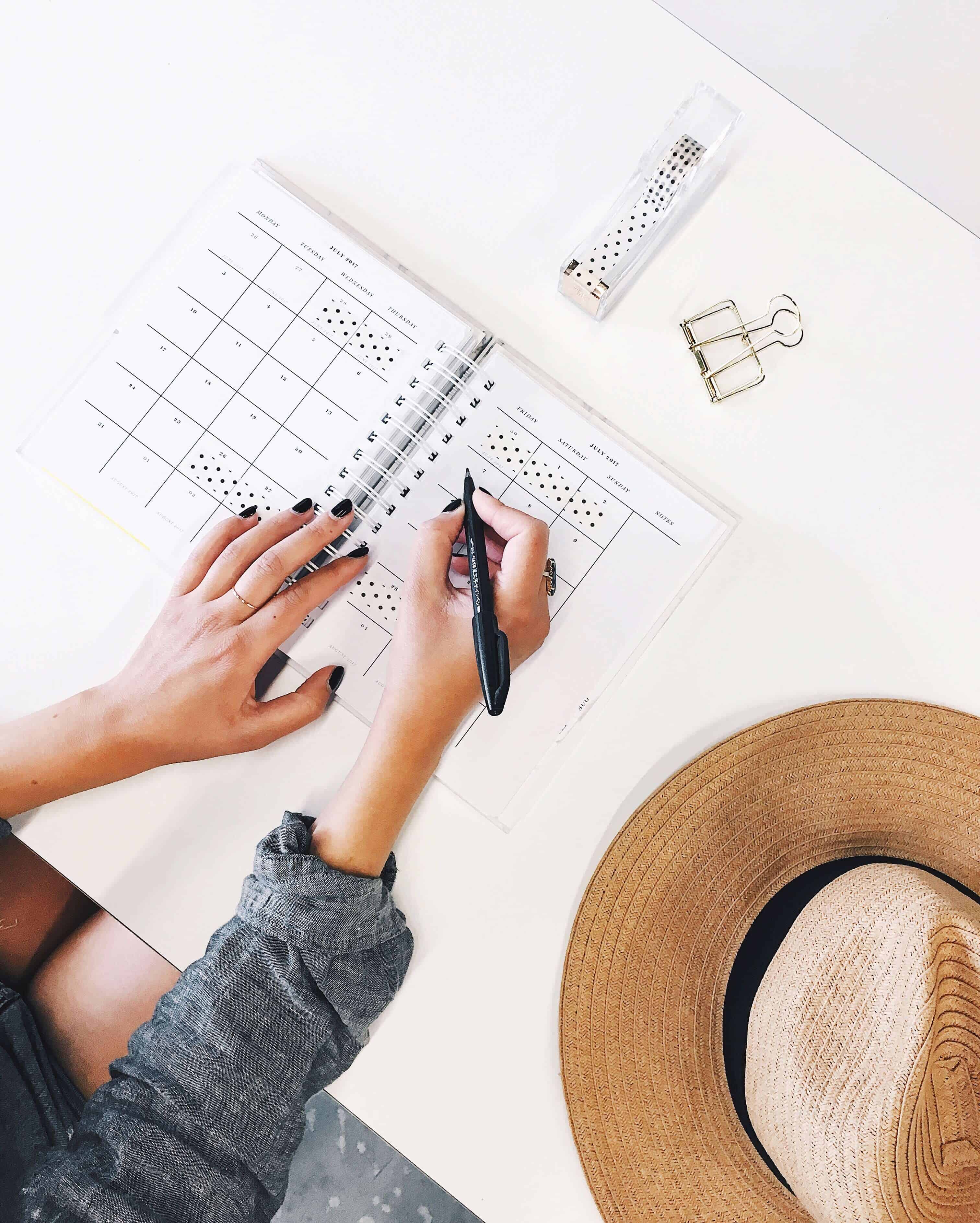 white desktop with woman writing in her planner, hat on desk