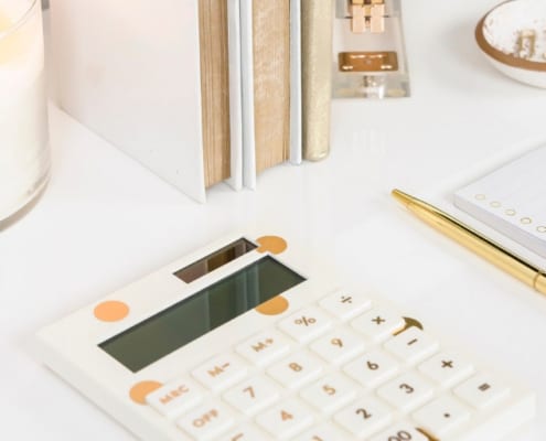 white desk with white and gold calculator, gold pen, binders, and a candle