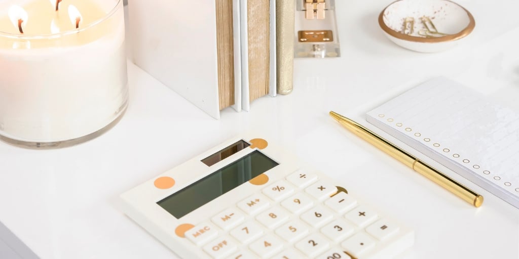 white desk with white and gold calculator, gold pen, binders, and a candle