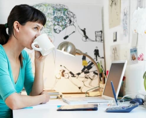 young woman drinking coffee at desk, looking out window thinking about best financial decisions