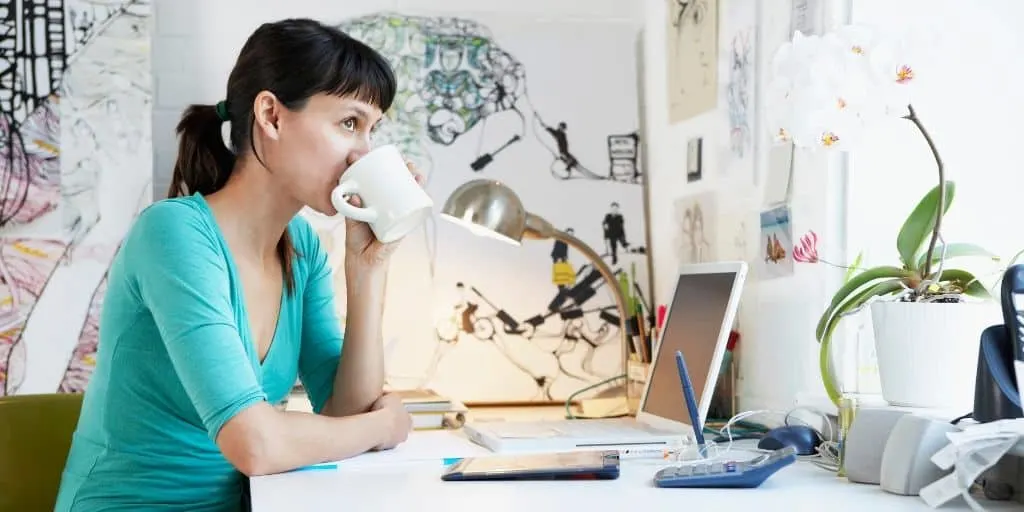 young woman drinking coffee at desk, looking out window thinking about best financial decisions