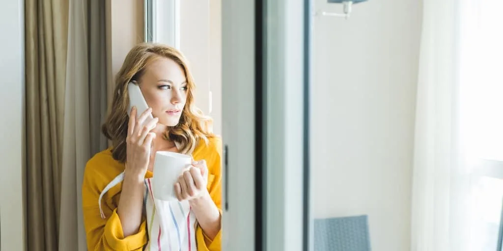 woman with yellow sweater on phone, looking anxiously out window, deal with debt collectors