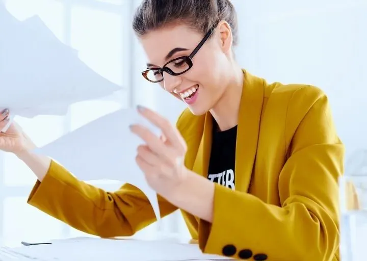 young professional woman in mustard business jacket, smiling at tons of paperwork