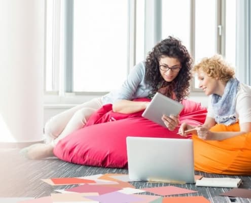 two women sitting on pink bean bag chair, looking at ipad screen together