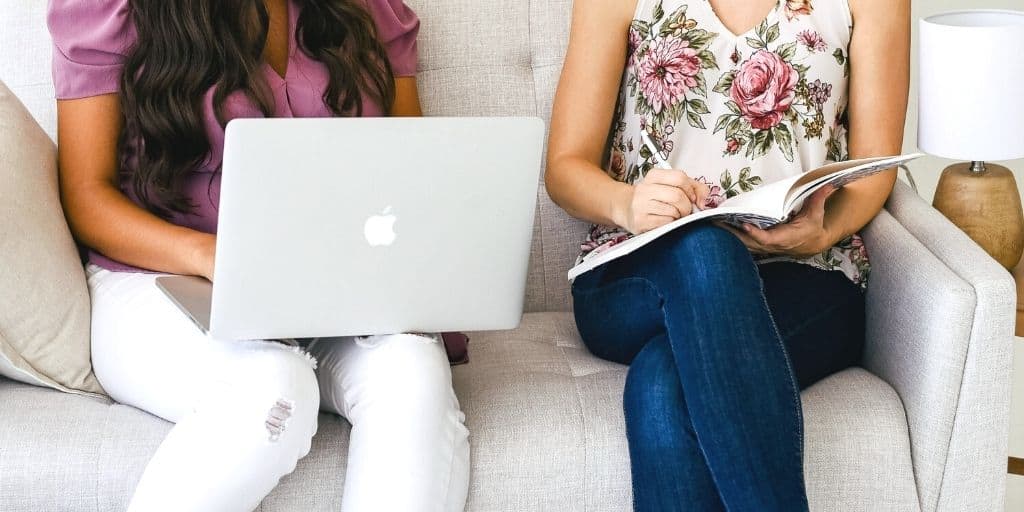 two women on sofa, one on laptop shopping online and wondering how to stop spending money online shopping