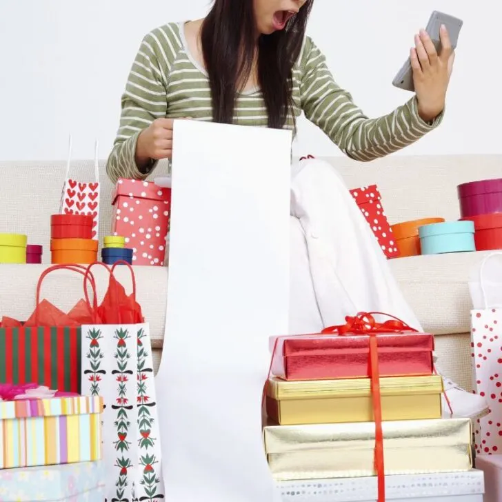 woman looking shocked at phone with long receipt piling over huge amount of gifts