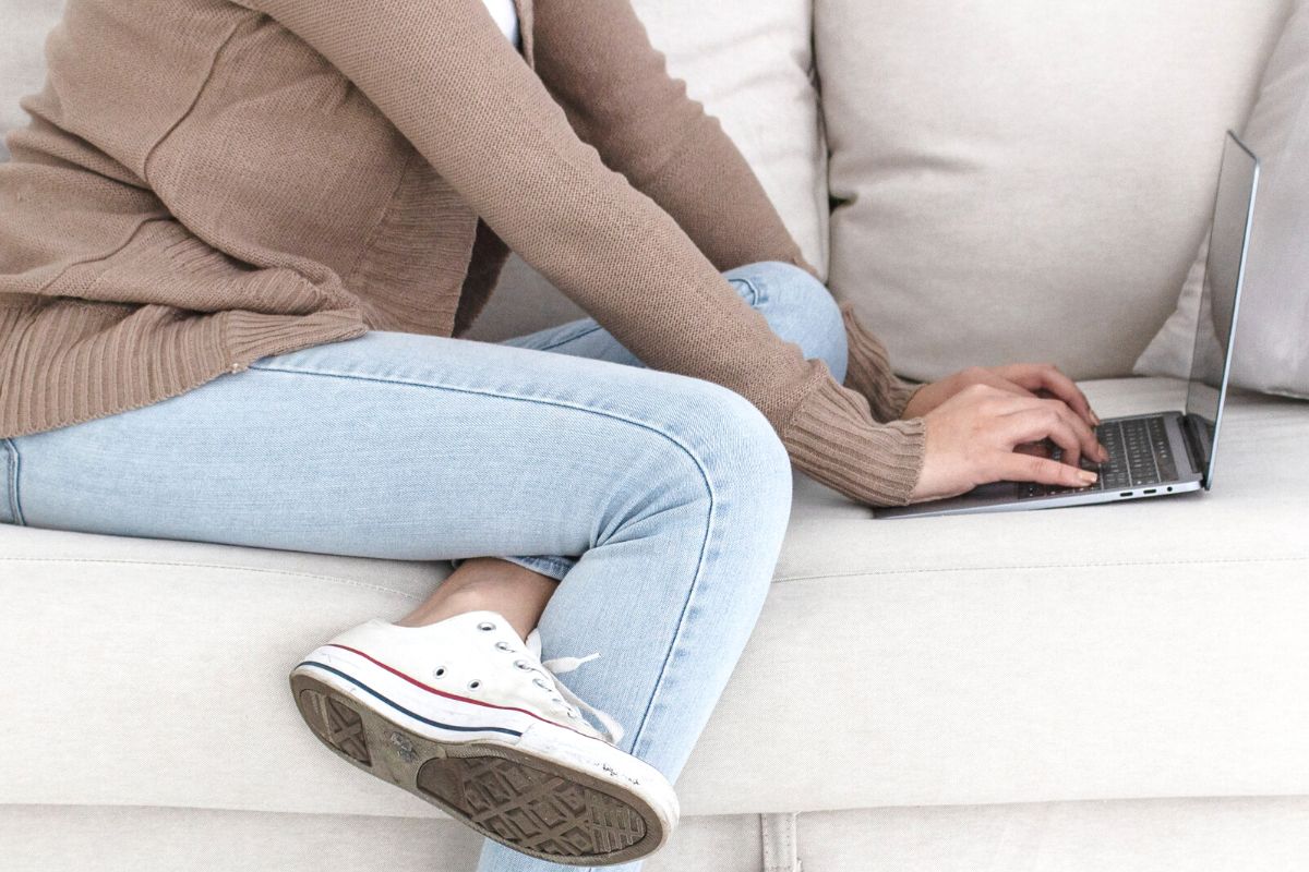 woman in sneakers on white couch, typing on computer