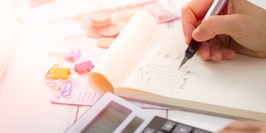 hand with pen, writing on a pad of paper, desk with calculator