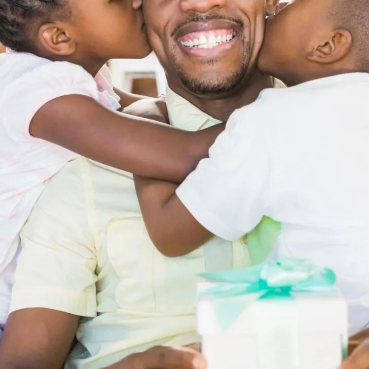 man getting kissed by two kids, smiling, with gift in lap