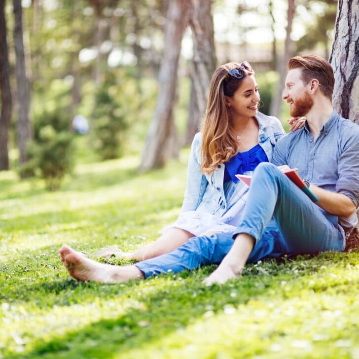 couple sitting up against tree in park reading something from a book and smiling