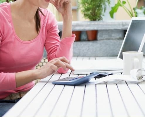 woman in pink shirt sitting in front of laptop with calculator, tired of living paycheck to paycheck
