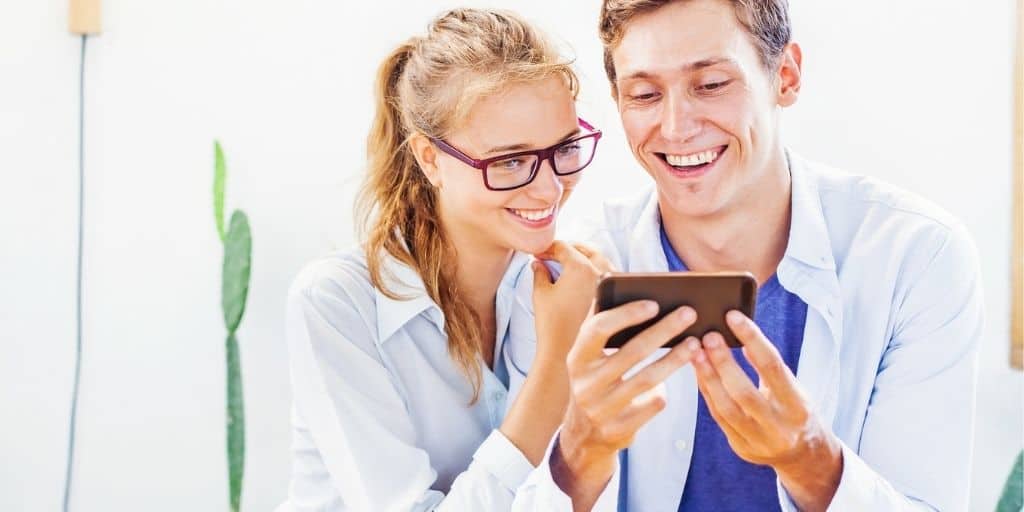 young couple sitting next to each other looking at best budget couple app on their phone together, smiling