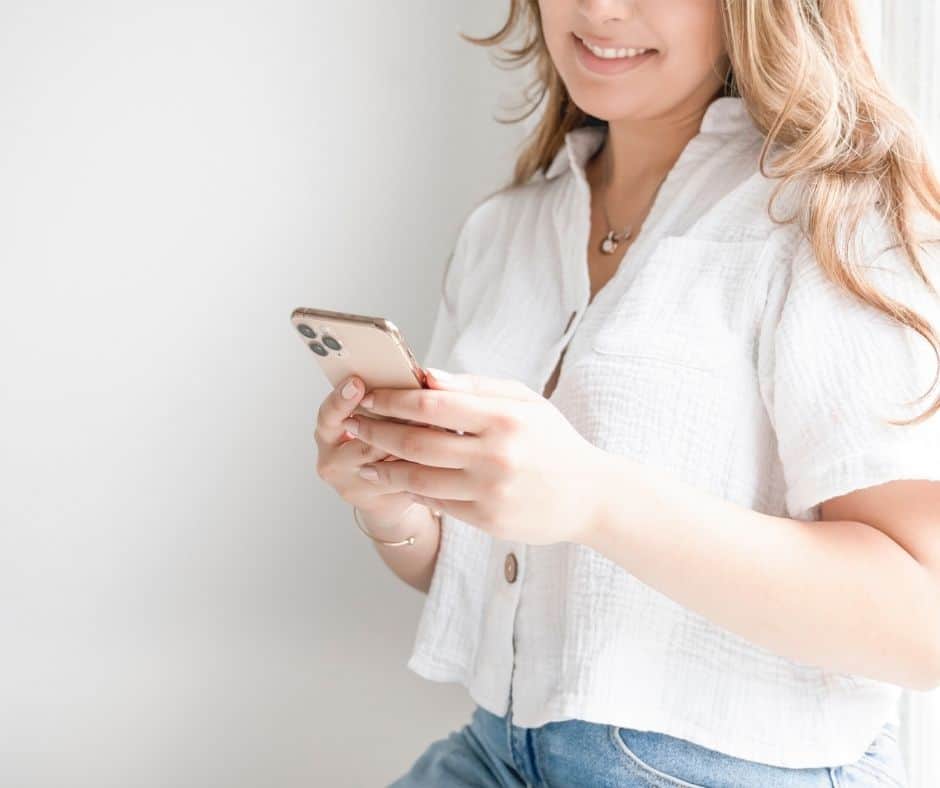 woman standing against wall, smiling, downloading money mindset app to phone for money mindset tips