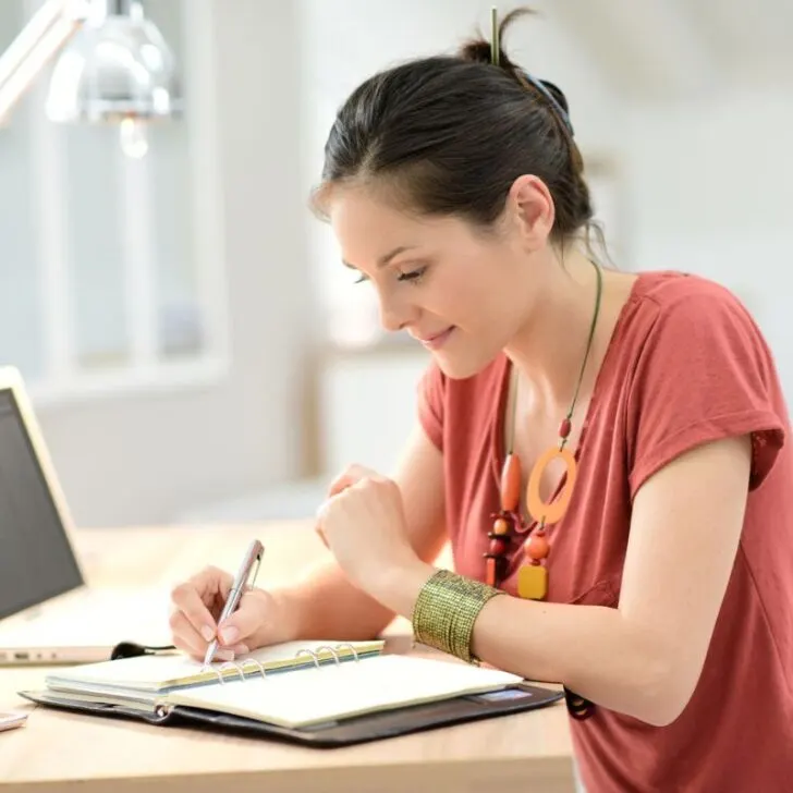 woman in orange shirt and messy bun working at home desk on spending less