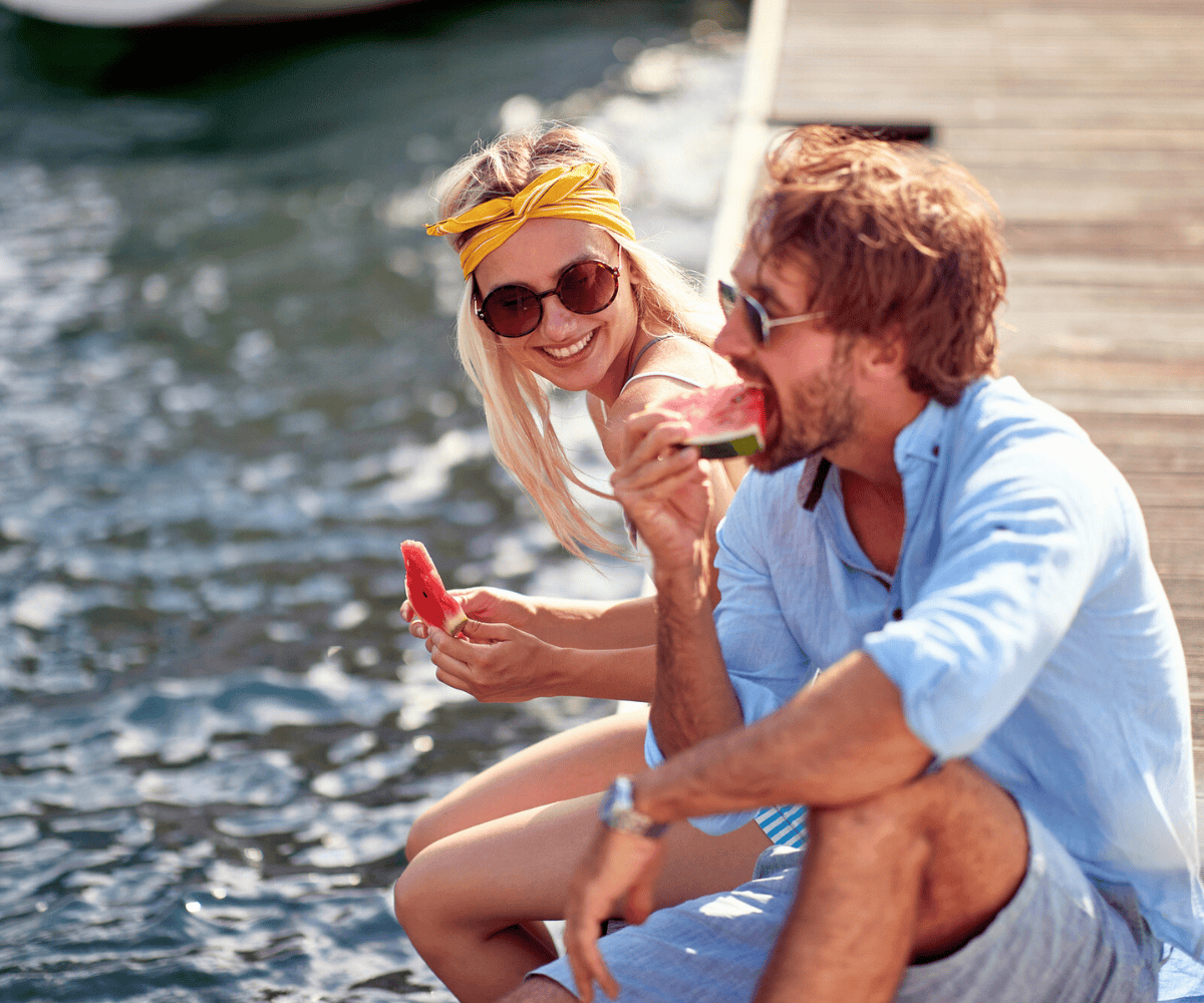 couple in sunglasses eating watermelon off the dock, dangling feet in water