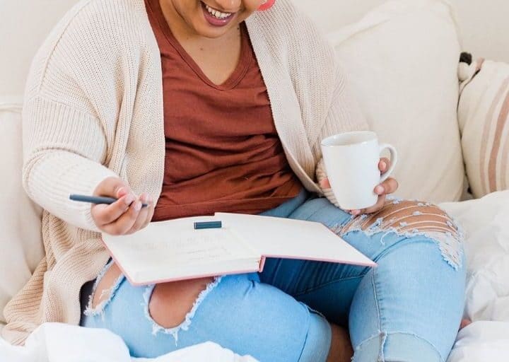 black woman filling out no spend tracker on bed with coffee, smiling