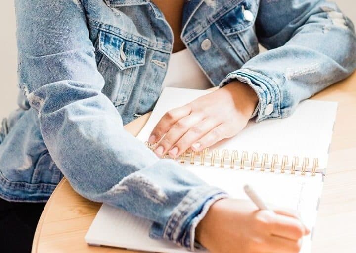 woman in jean jacket at wooden desk filling in a budget sheet notepad