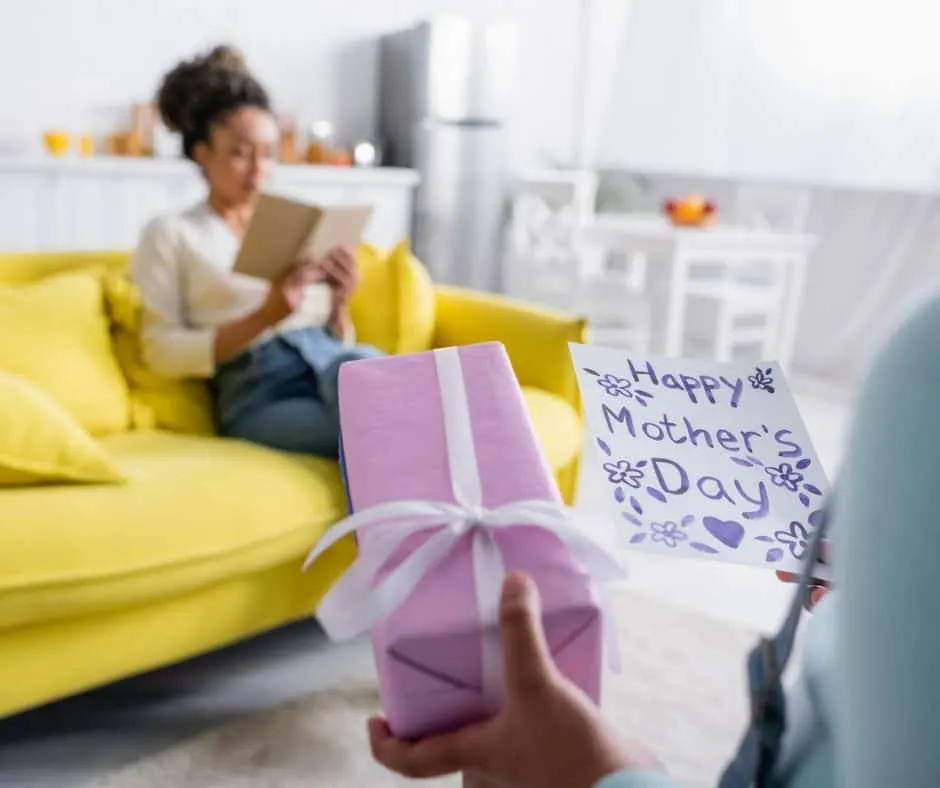 child surprising unsuspecting mother sitting on couch reading with purple-wrapped gift, for gifts for mom who has everything