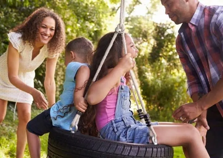 family of four laughing and playing on a tire swing out back as a way to cut family expenses