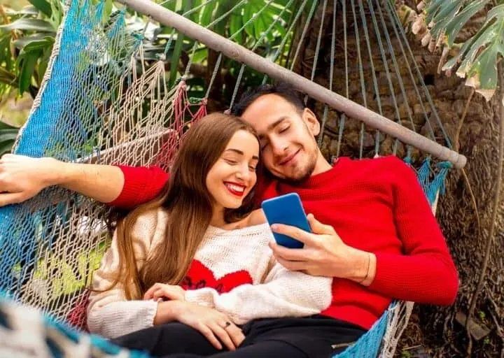 young couple in red and white shirts on a hammock, smiling, looking at summer couple activities on their phone