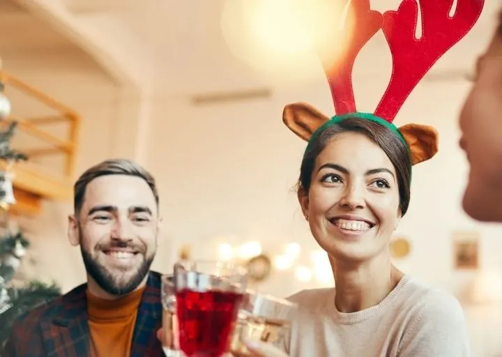 man and woman with Rudolph antlers in front of tree, smiling while doing adult Christmas Eve traditions