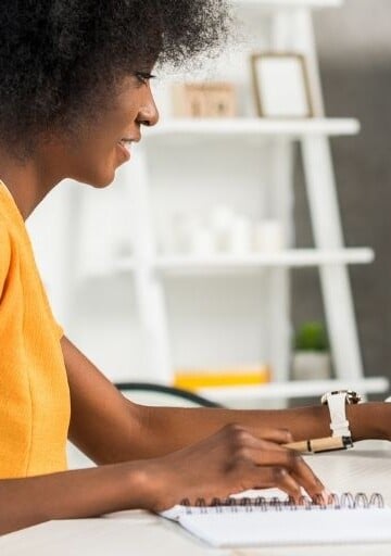young woman in yellow shirt at desk, thinking about which financial goal category she wants to go after