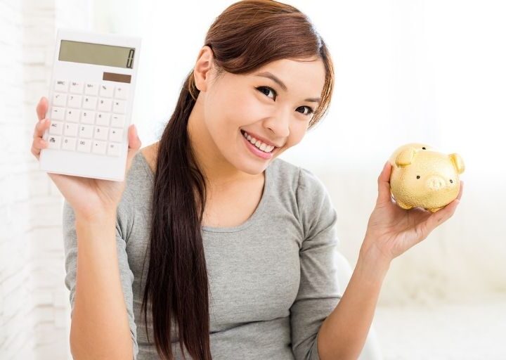 young woman smiling, holding calculator and piggy bank to track her money saving goal