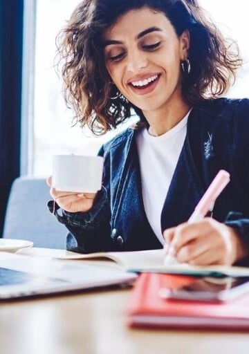 middle aged woman smiling with coffee working on steps to achieve savings goal at desk