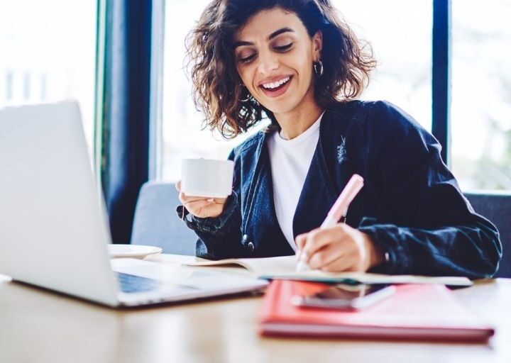 middle aged woman smiling with coffee working on steps to achieve savings goal at desk