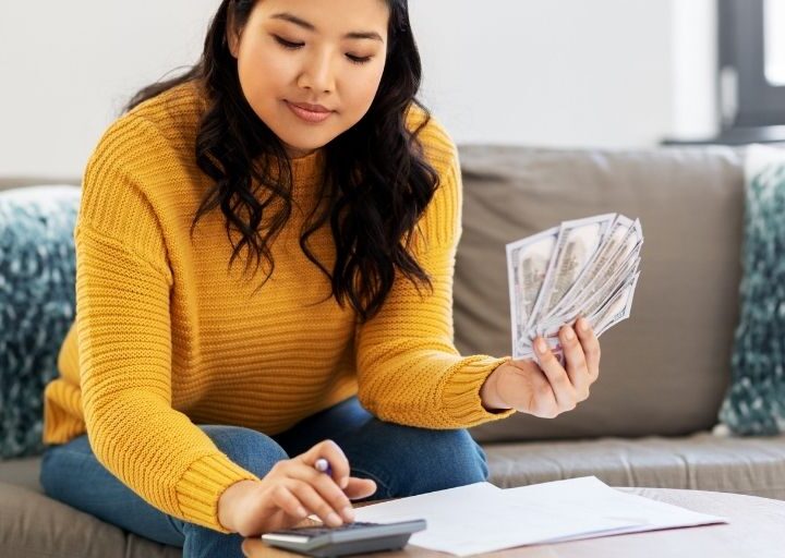 young woman in orange sweater with money calculating daily money saving challenge amount