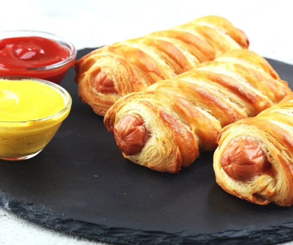 three pigs in a blanket on slate plate with mustard and ketchup
