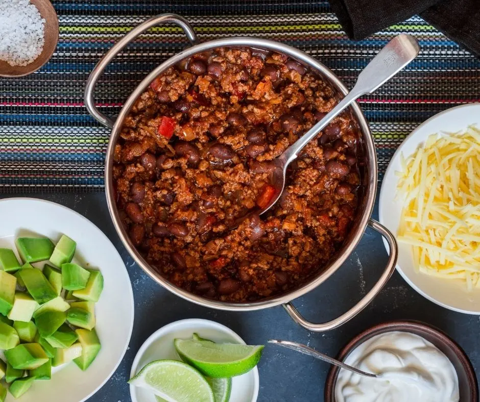 pot of chili in center with cubed avocado, cheese, onions, and limes