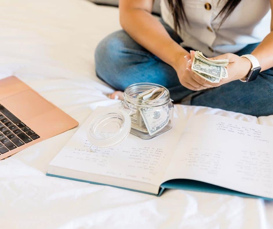woman with jar of money on bed, notebook and laptop figuring out her frugal living tips strategy for a big impact