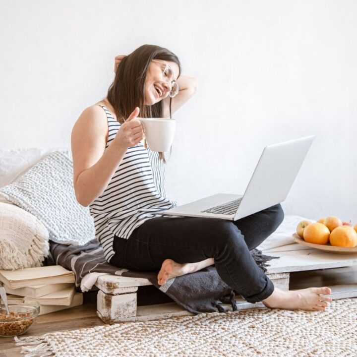 woman with coffee cup on laptop, on pallet bed, smiling, reading about benefits of frugal living