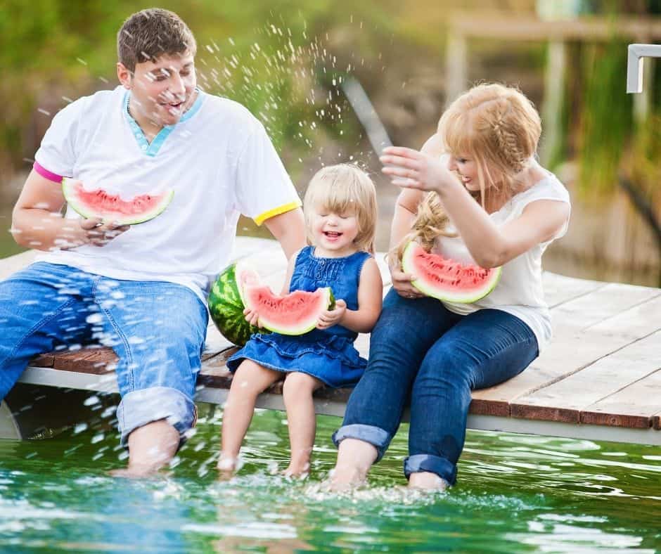 young family sitting on dock eating watermelon, splashing water, and having a good time
