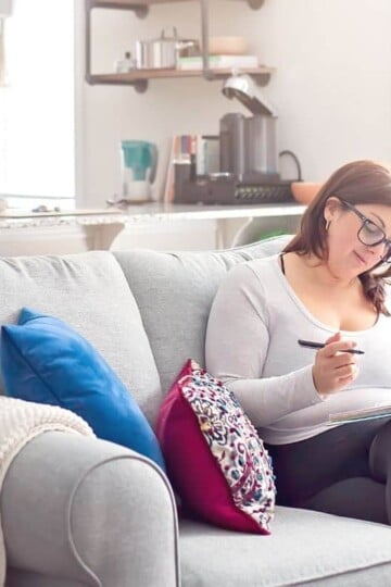 woman in glasses on couch, working on writing out her savings challenge