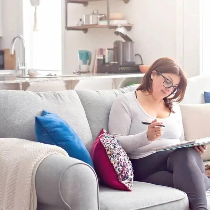 woman in glasses on couch, working on writing out her savings challenge