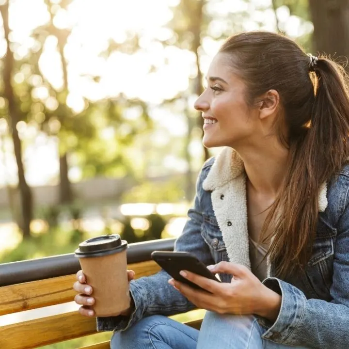 woman sitting on park bench with laptop and coffee, smiling at fall foliage