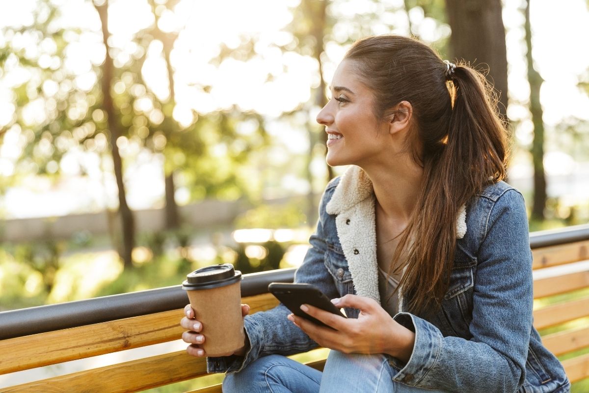 woman sitting on park bench with laptop and coffee, smiling at fall foliage