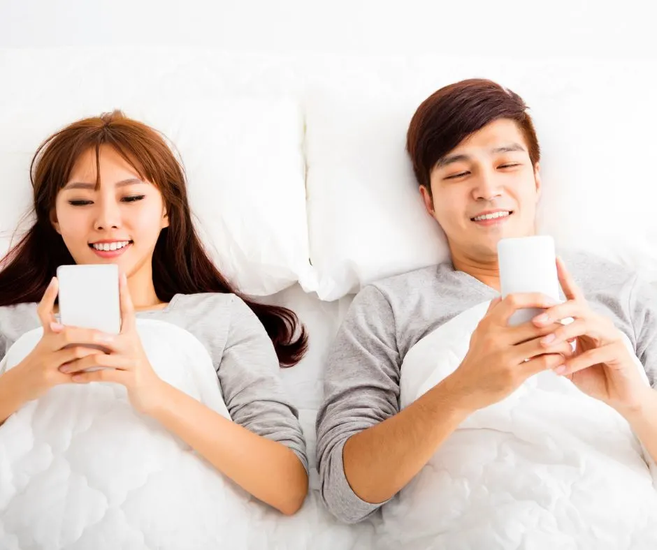 couple in white bed, looking at intimate phone apps for at home date idea and smiling