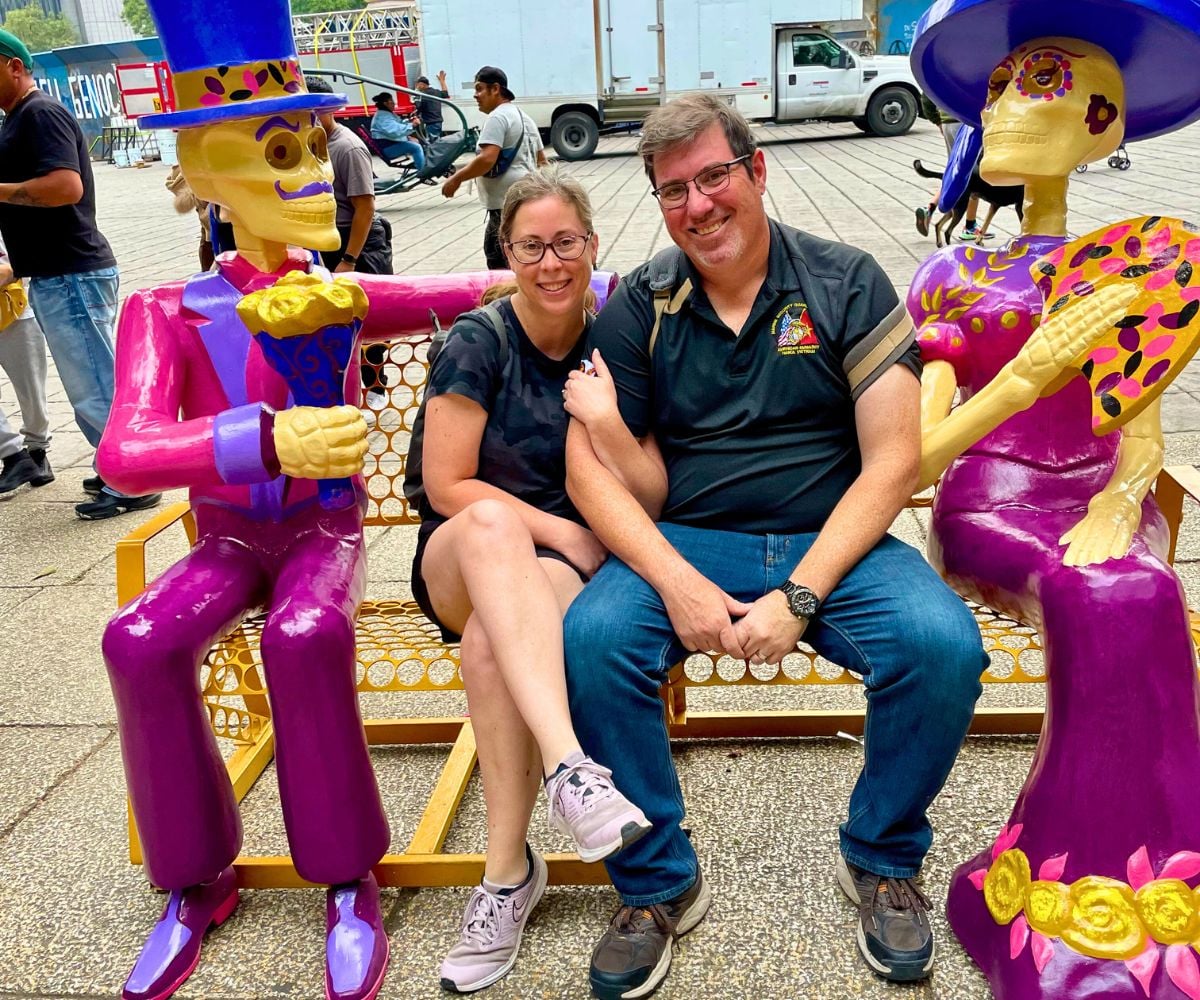 author with husband in Mexico City sitting in between two man/woman Day of the Dead skeletons on bench