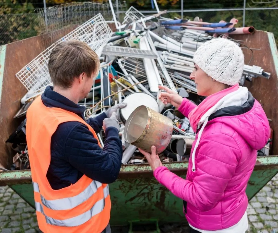 woman in pink coat at container filled with scrap metal, talking to man about selling scrap metal