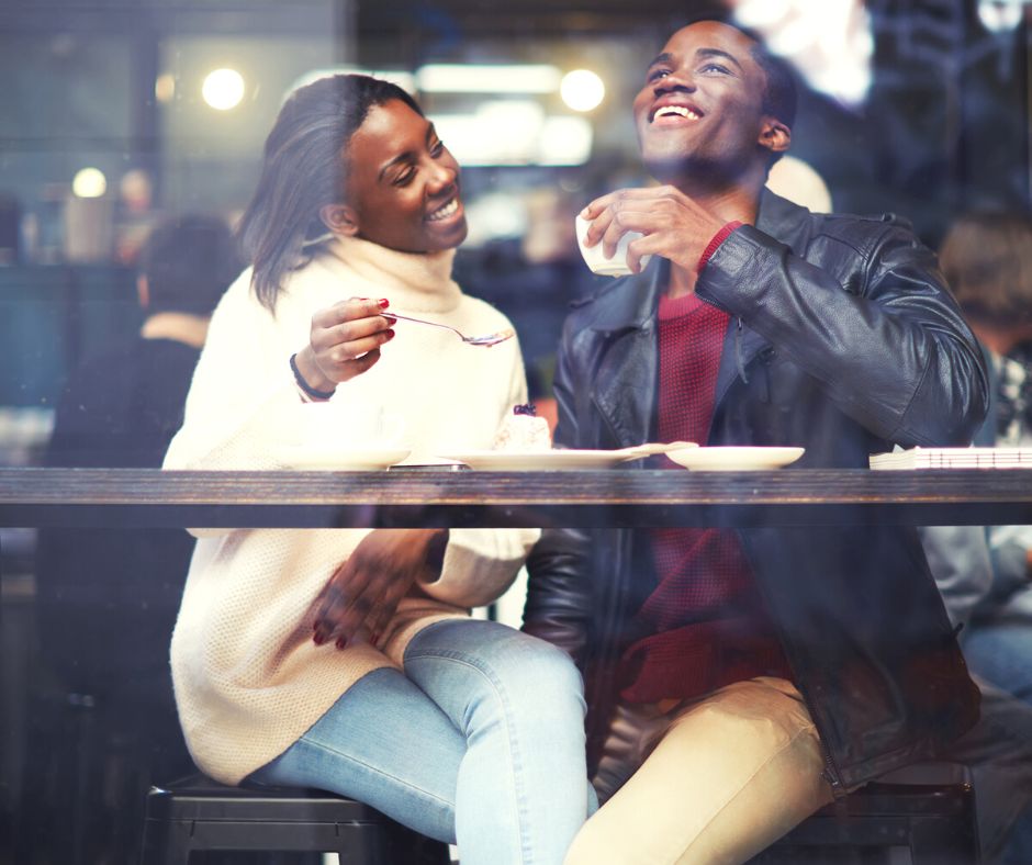 couple in coffee shop window, laughing and eating cake together on coffee shop date