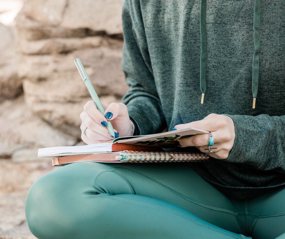 woman with dark teal nail polish and green jogging suit sitting on rocks and writing in gratitude journal