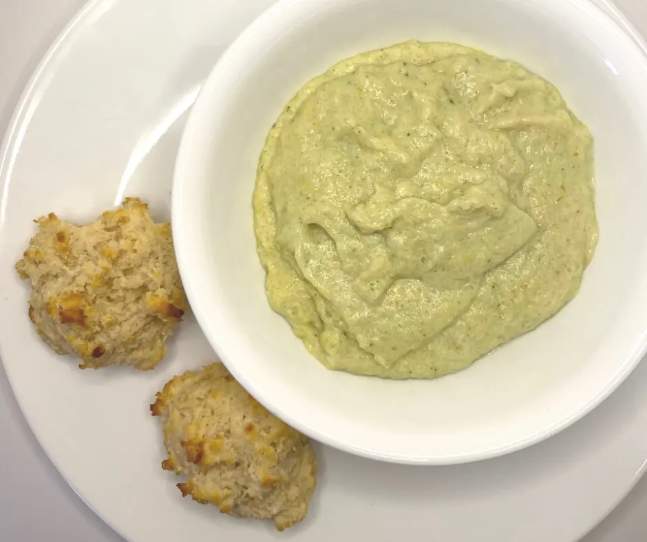white bowl of pureed broccoli soup with two cheddar biscuits on side on white plate
